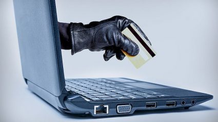 ręka w czarnej rękawiczce z kartą płatniczą, wyłaniajaca się z ekranu laptopa,