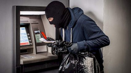 przestępca w kominiarce przy bankomacie, z kartą płatniczą w dłoni