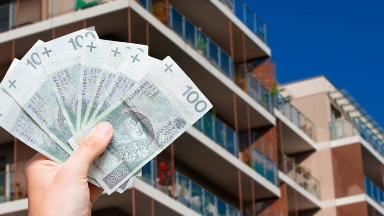 dłoń z plikiem banknotów na tle budynku mieszkalnego